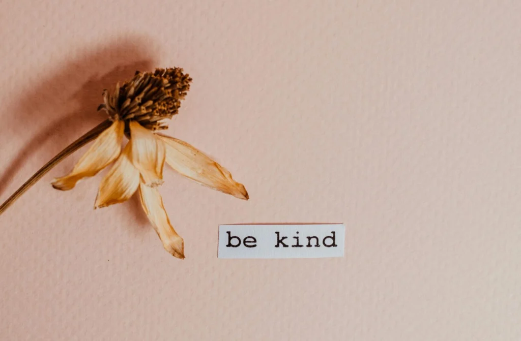 A minimalist image featuring a dry flower next to 'be kind' text on textured brown paper.